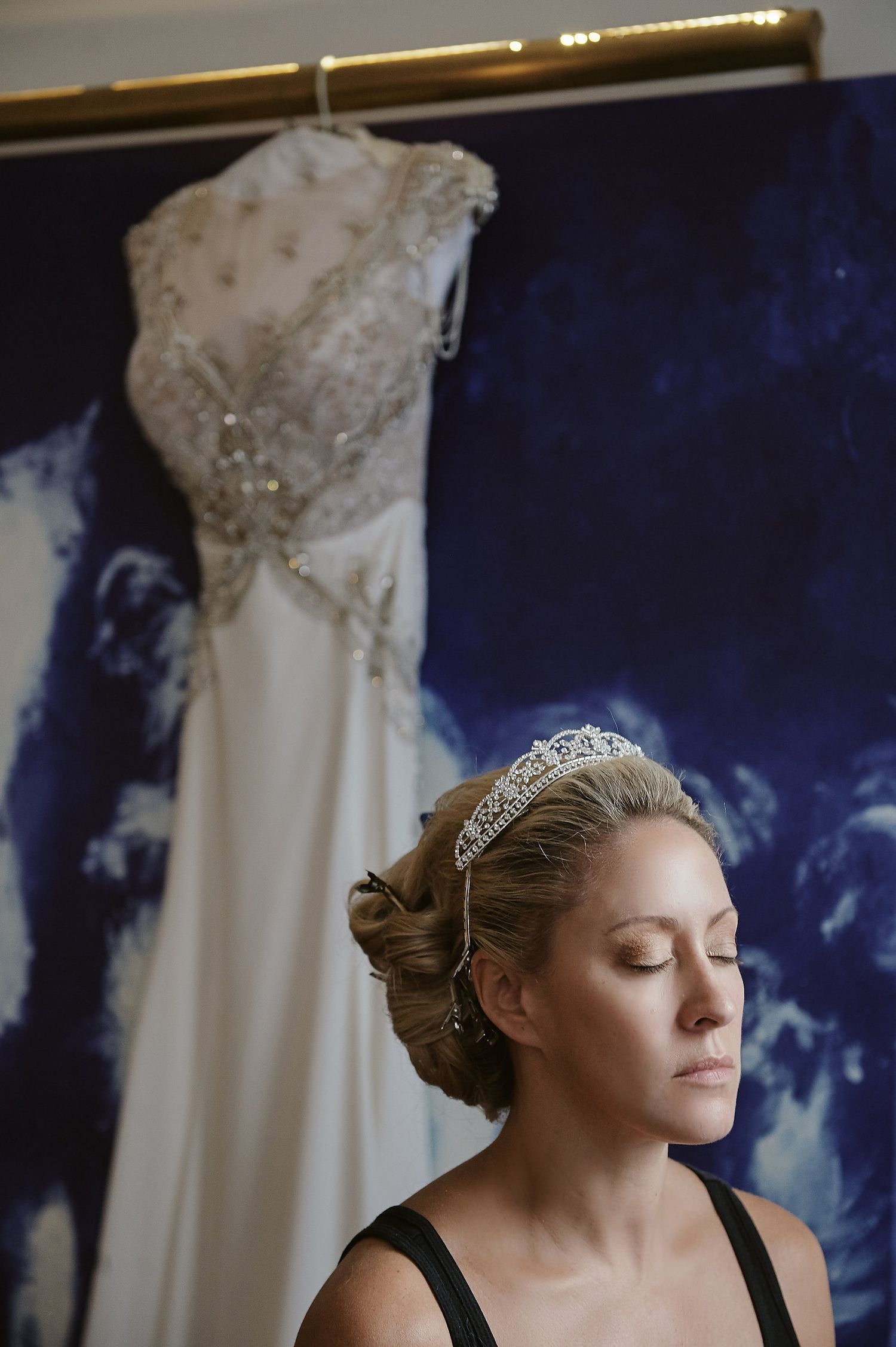 An elegant white beaded wedding dress hangs in the background against a dark blue backdrop.