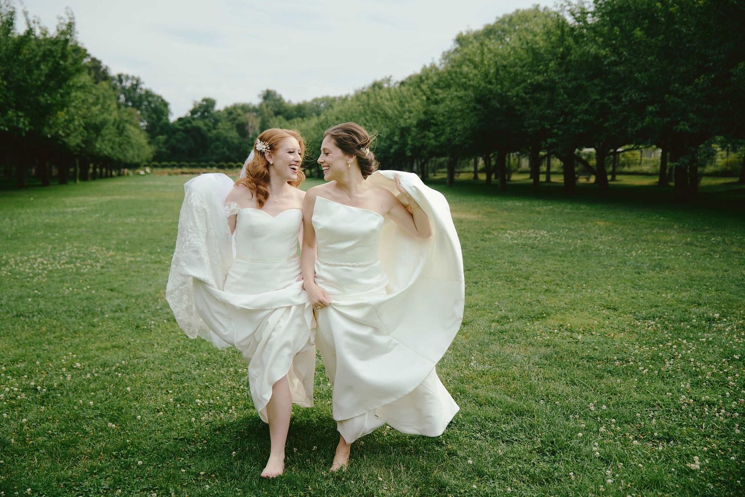 Two people in flowy white dresses run playfully across a green lawn surrounded by trees.