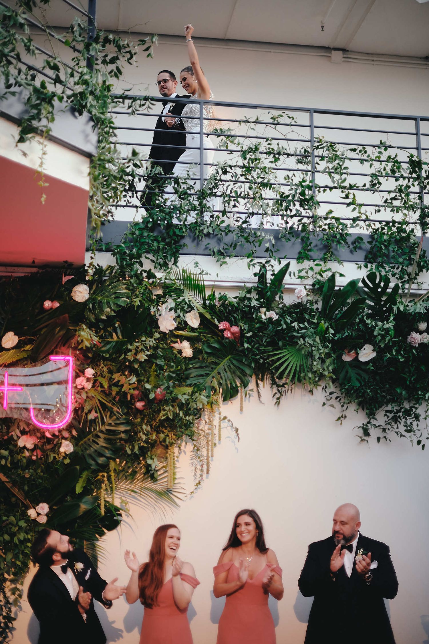 Wedding party members applauding beneath a decorated indoor floral installation with greenery and pink blooms.