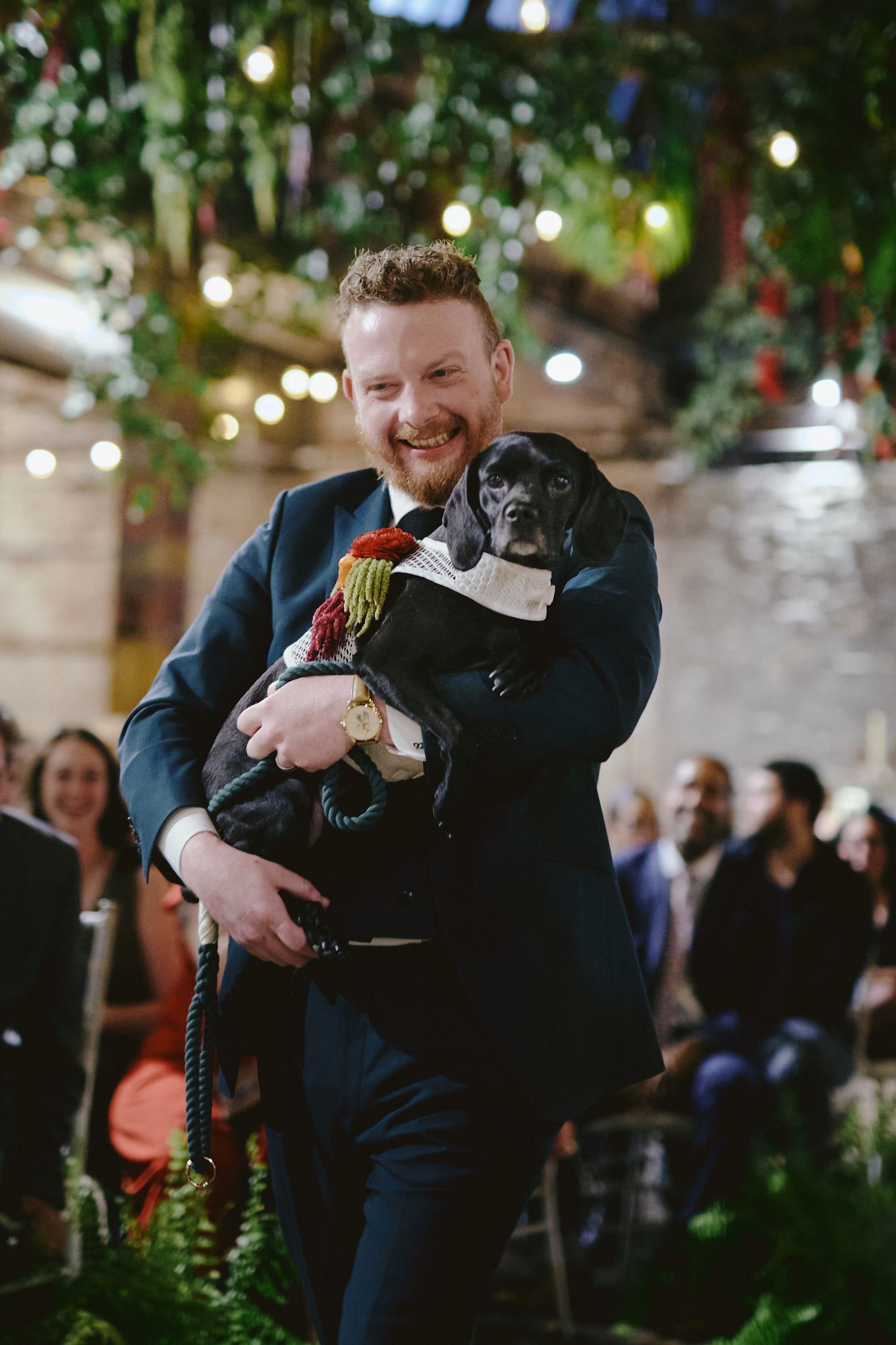 A black pug wearing a bow tie is held lovingly at an indoor wedding ceremony with twinkling lights and greenery.