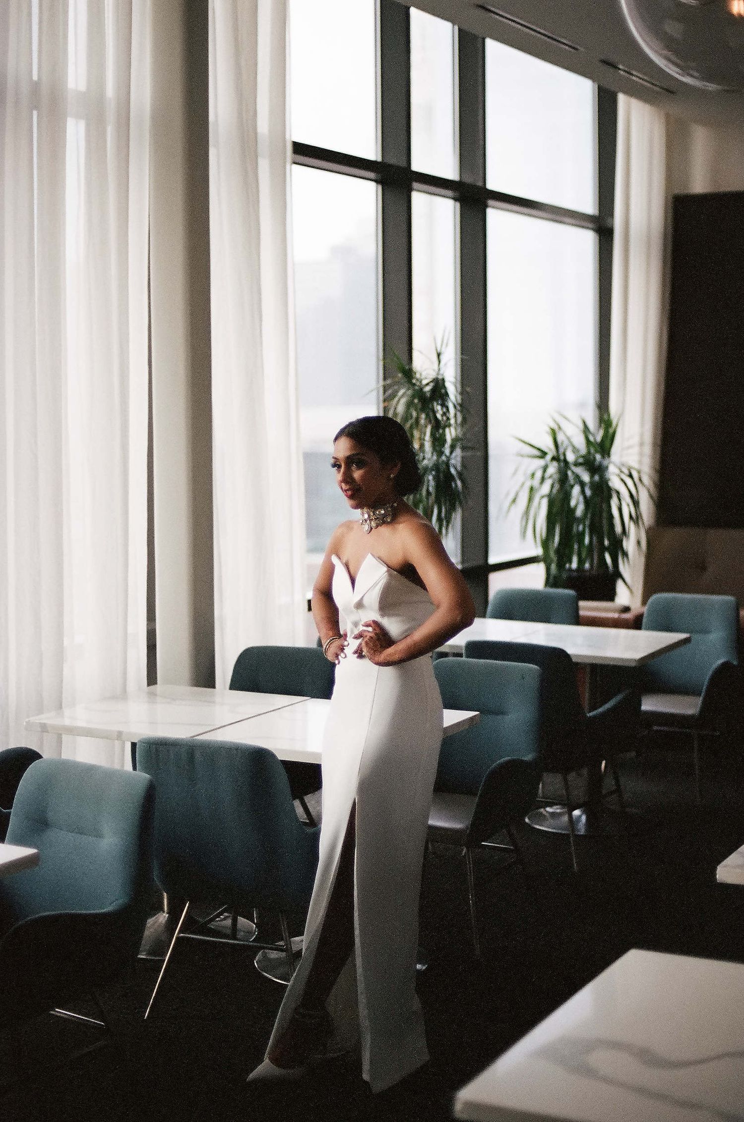 Elegant white wedding gown silhouetted against large windows in modern dining space with blue chairs and natural light.