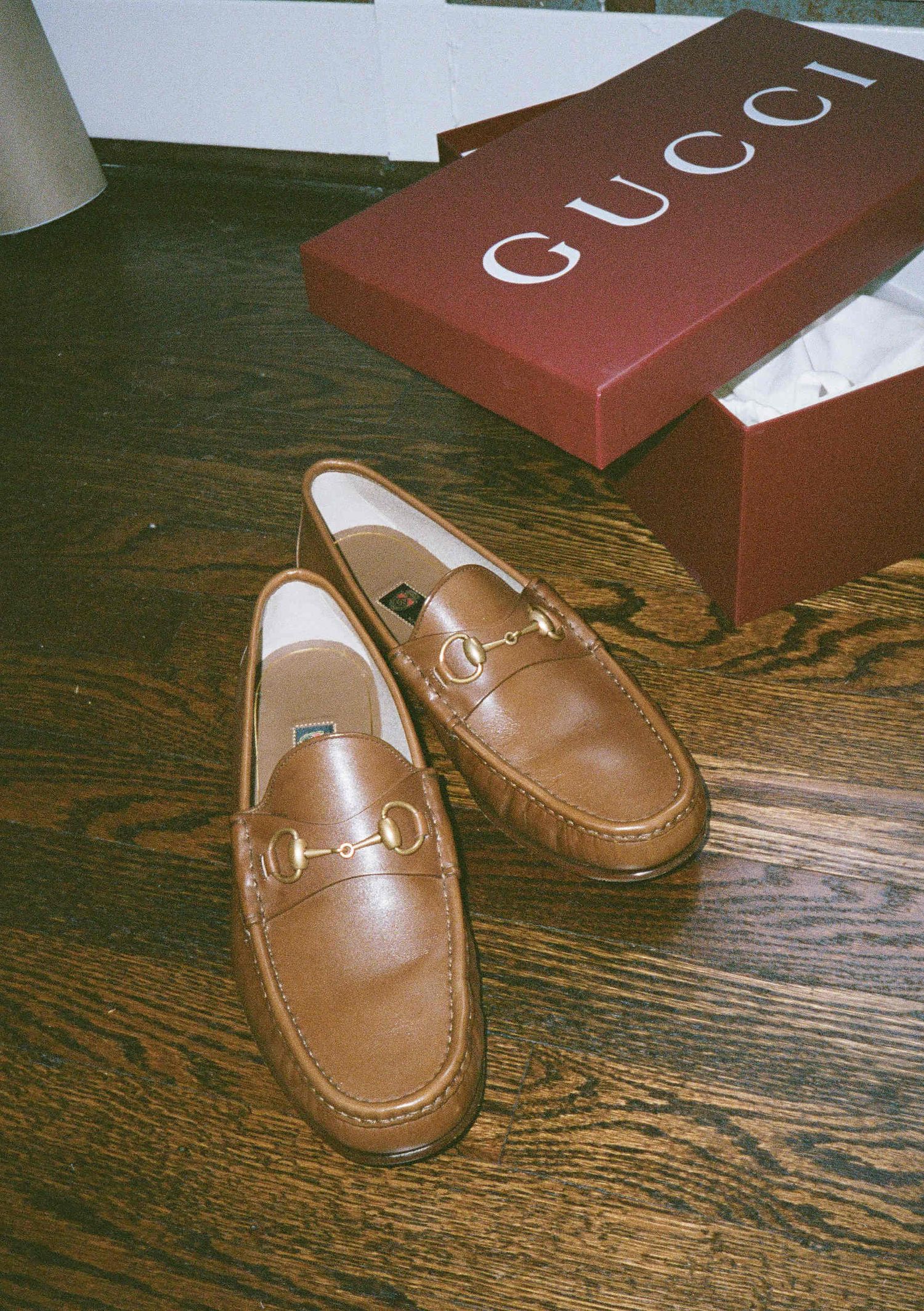 Brown leather Gucci loafers displayed on wooden floor with signature red Gucci box in the background.
