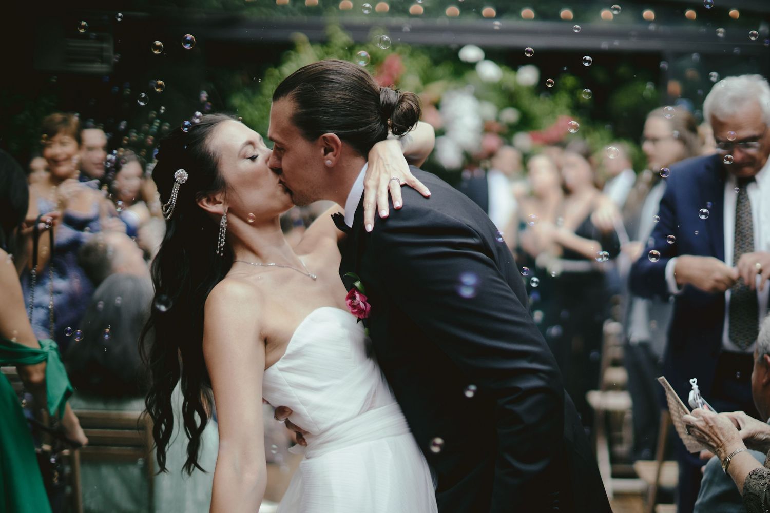 A romantic kiss at an outdoor wedding ceremony while wedding guests applaud and celebrate the joyful moment.