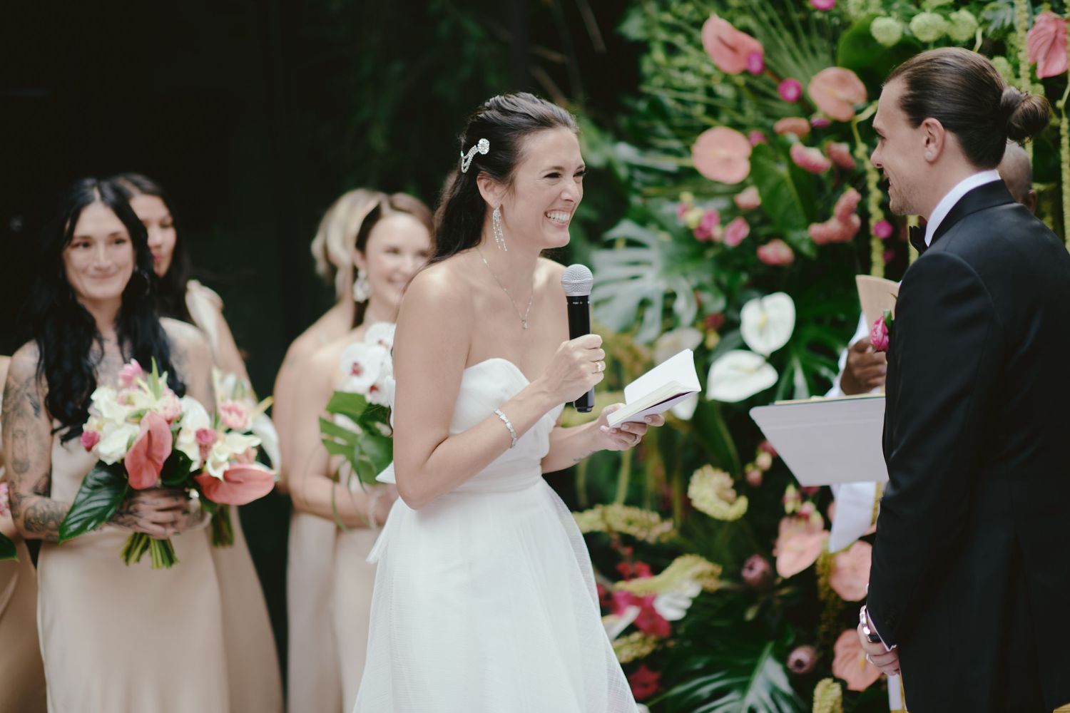 A joyful outdoor wedding ceremony with bridesmaids in beige dresses and a floral arch backdrop.