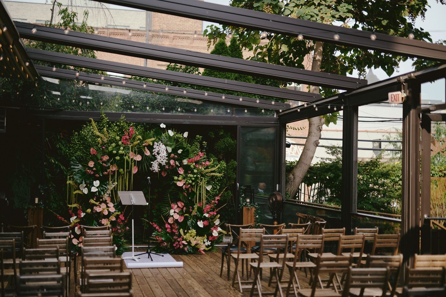 Rustic outdoor wedding ceremony setup with wooden chairs under a glass-covered pergola decorated with floral arrangements.