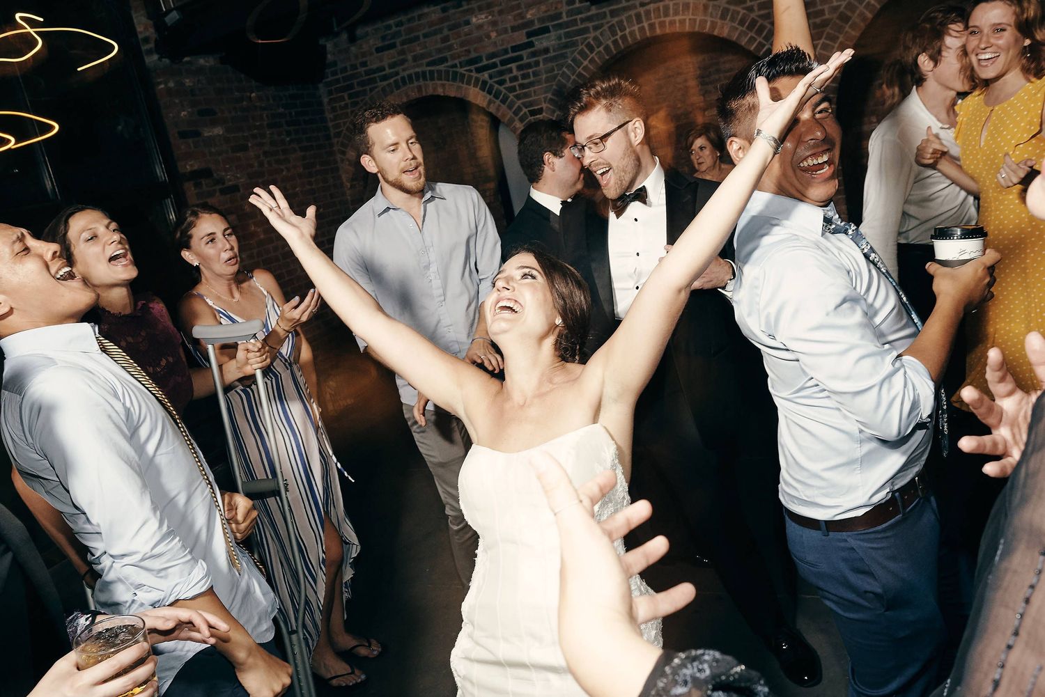 Wedding guests dancing and celebrating with arms raised in a brick-walled reception venue.
