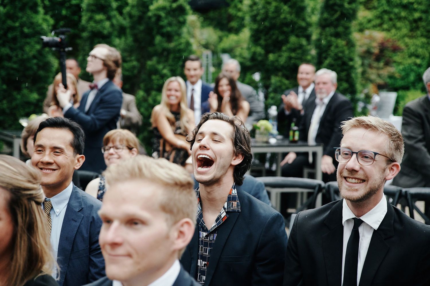 Guests react with joyful laughter while seated at an outdoor wedding ceremony.