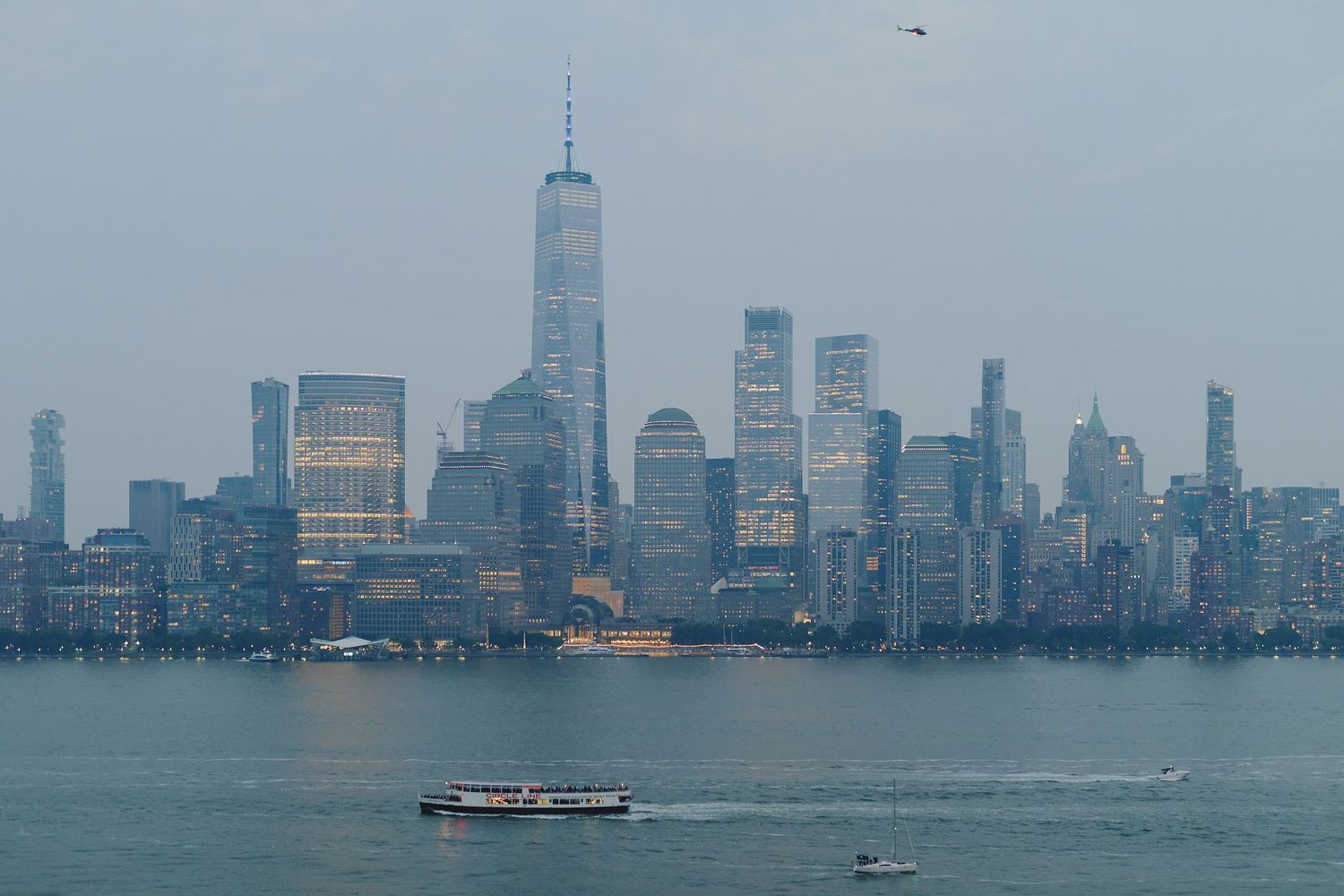 Manhattan skyline with Freedom Tower rises above New York Harbor as boats cruise across foggy waters at dusk.