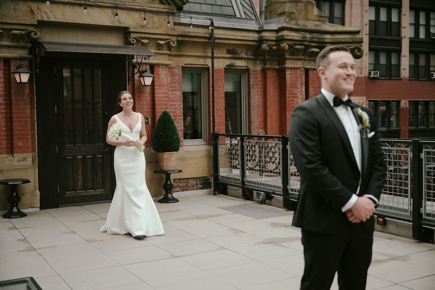 A wedding first look moment between a bride and groom on a brick brownstone balcony in formal attire.