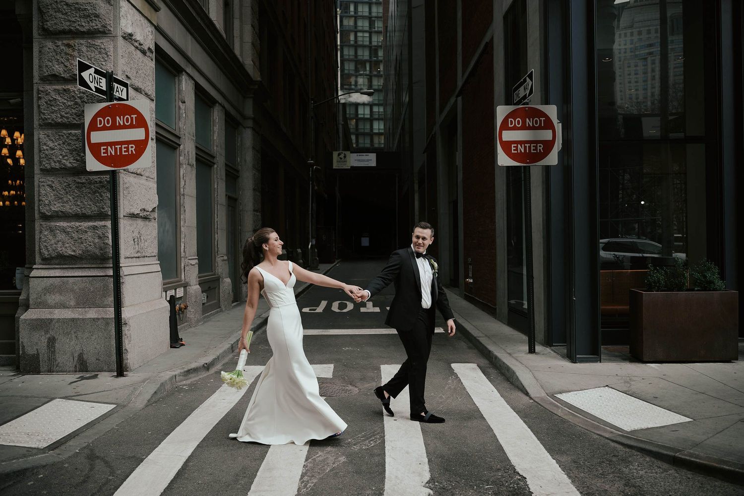 A bride and groom cross a city intersection at a crosswalk between two do not enter signs in an urban setting.