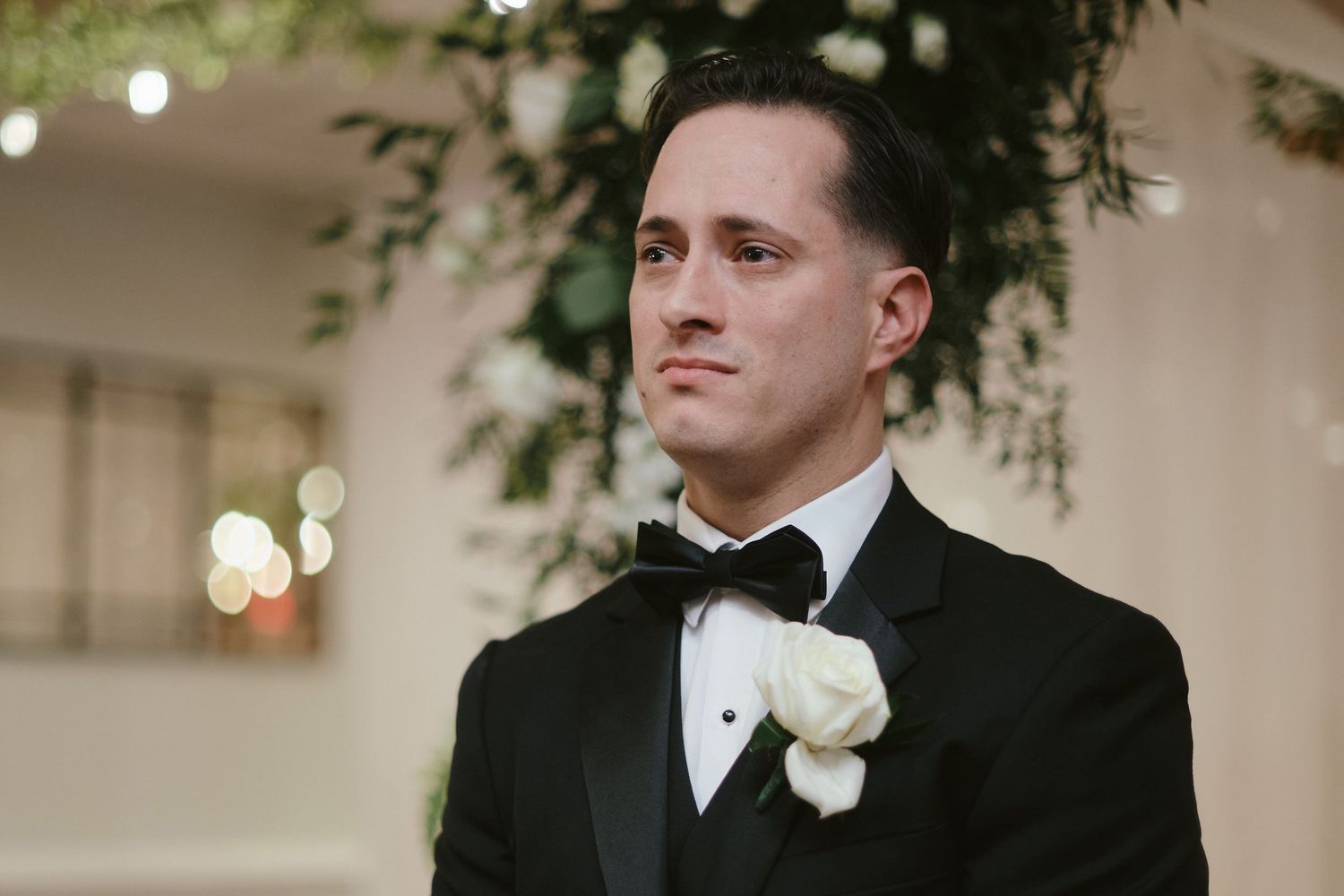 Groom wearing classic black tuxedo with white boutonniere stands in elegant indoor venue with bokeh lights.