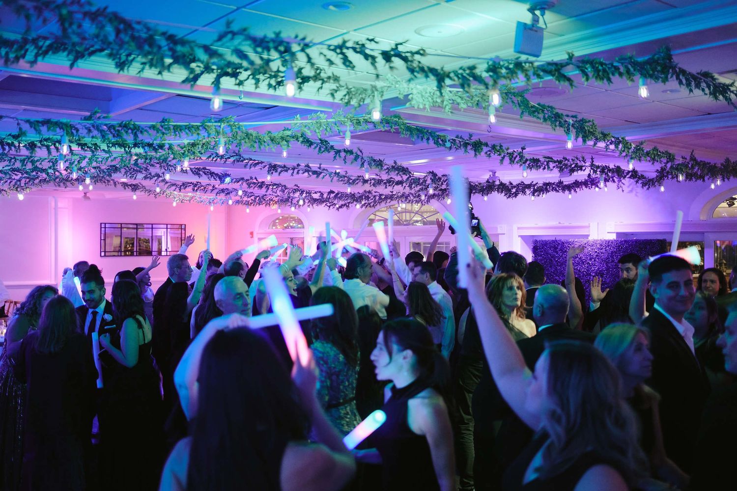 Crowd dances under purple and blue lighting with hanging twinkle lights at a lively reception party.