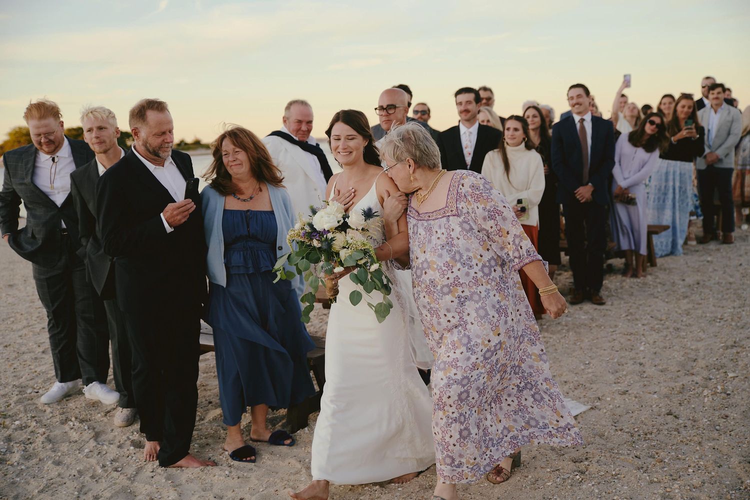 A wedding party walks along a sandy beach path at sunset with guests following behind in a long procession.
