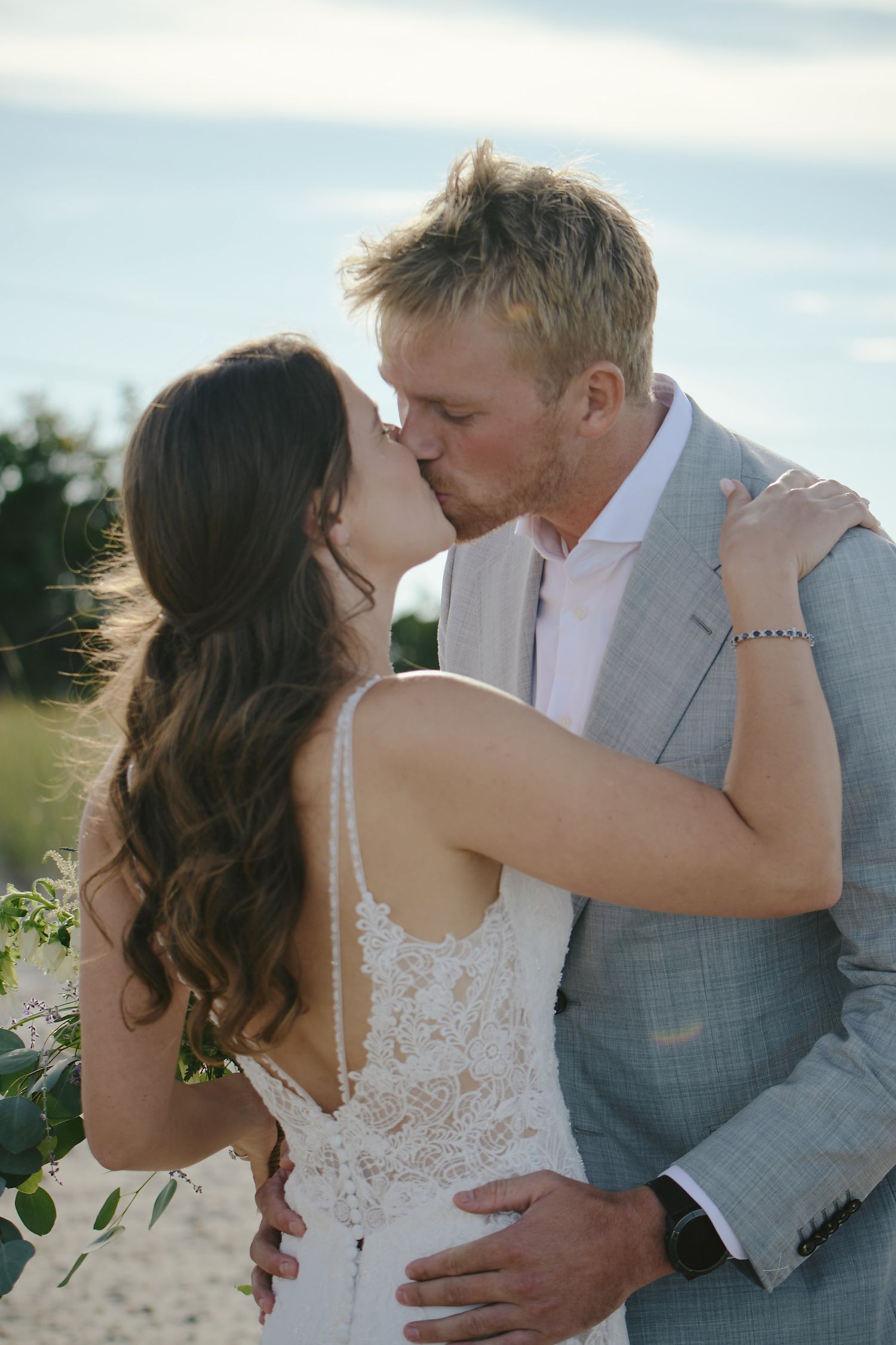A romantic wedding couple share a kiss on a sunny beach, with her wearing a lacy white dress and him in a gray suit.