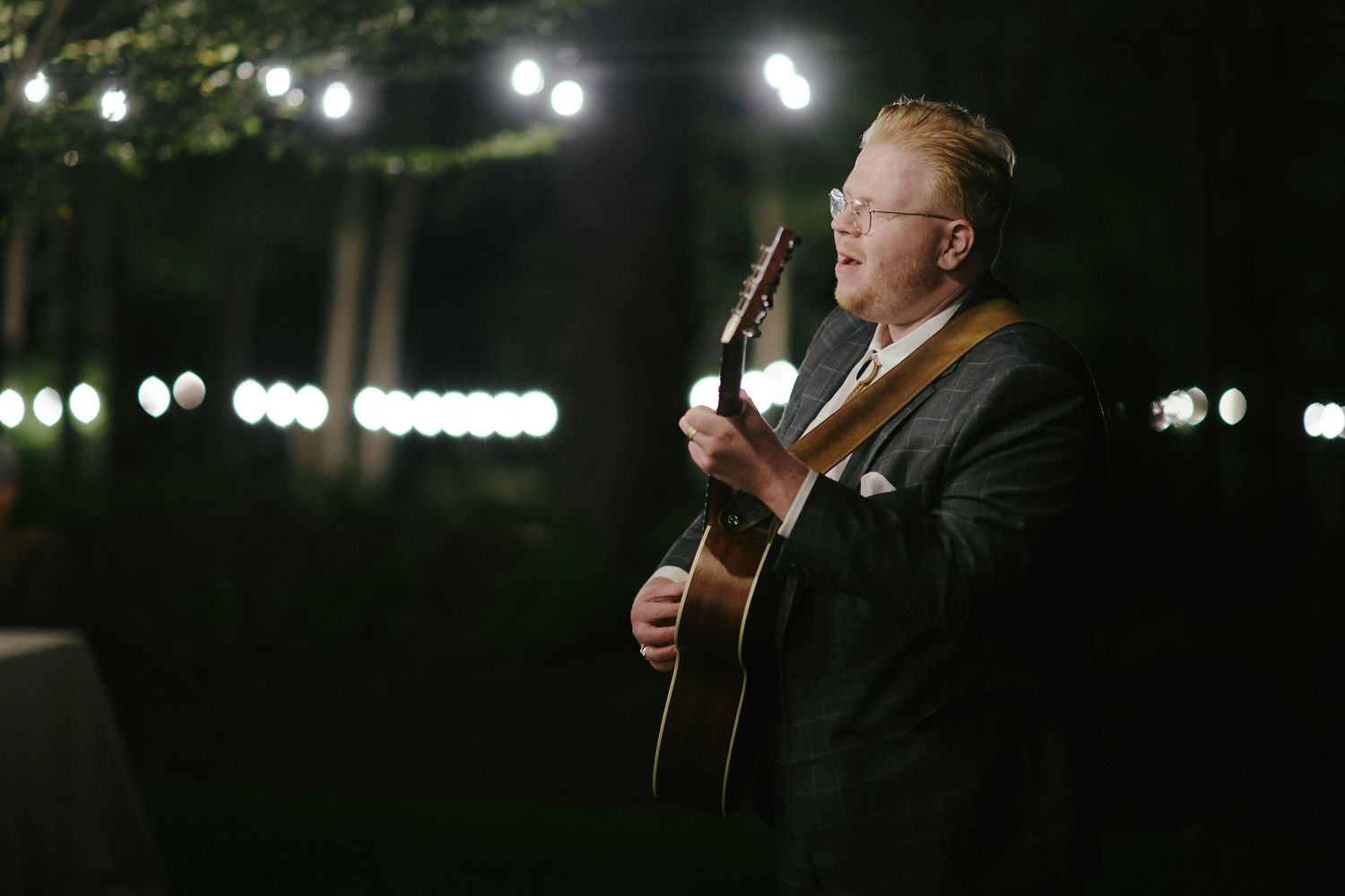 A person performs with an acoustic guitar on a dimly lit stage with glowing lights in the background at night.
