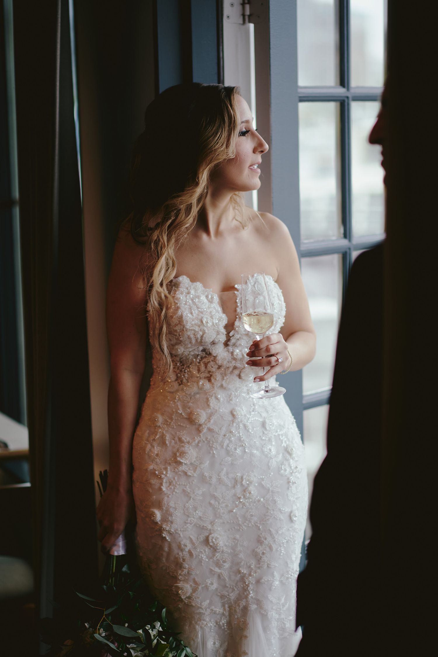 A person in an elegant lace wedding dress holds a wine glass while standing by a window in moody lighting.