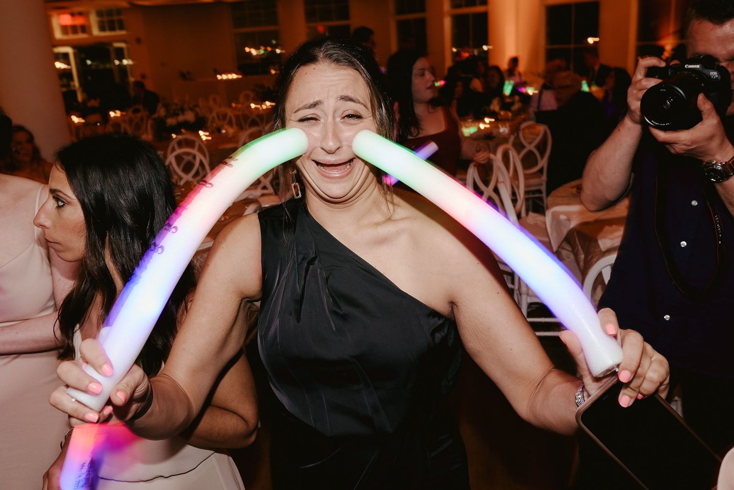 Person in black one-shoulder dress holds glowing LED foam light stick while dancing at crowded nighttime event.