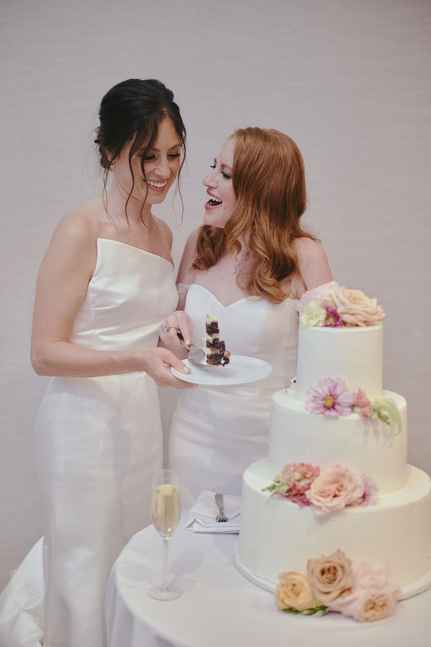 Two brides share a joyful moment with champagne and a three-tiered wedding cake decorated with pink and peach roses.