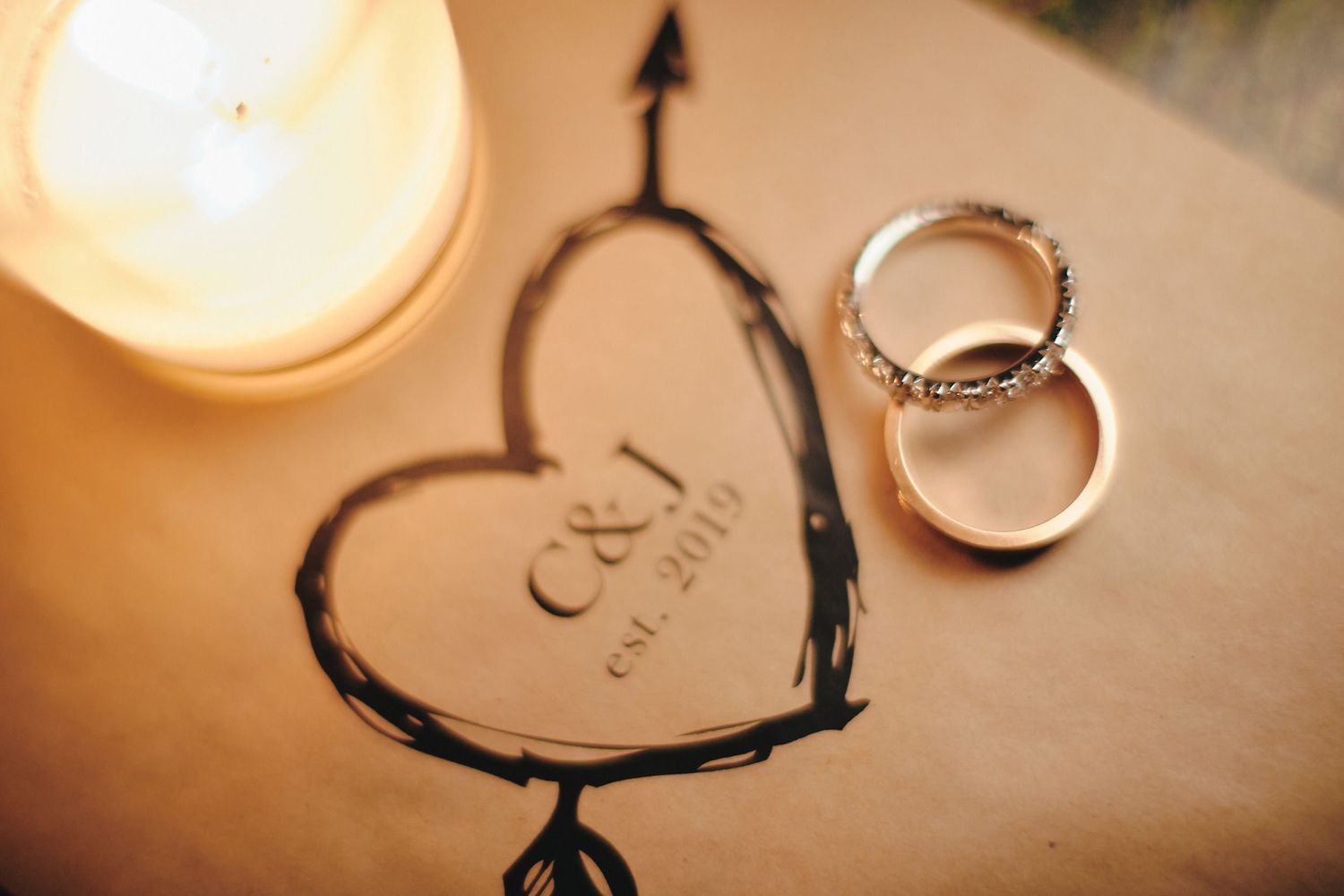 Wedding rings rest beside a hand-drawn heart with initials and a glowing candle in warm lighting.