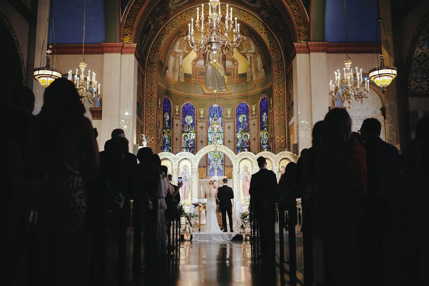 Silhouettes of wedding guests in a grand cathedral with ornate stained glass windows and crystal chandeliers.