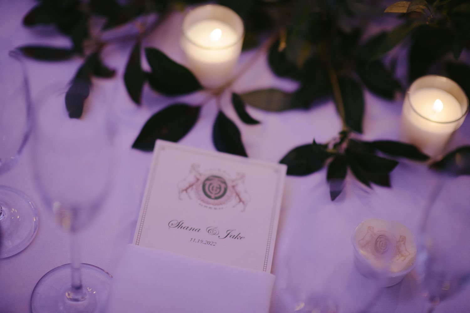 An elegant wedding reception table setting with illuminated candles and green leaves against a soft purple background.