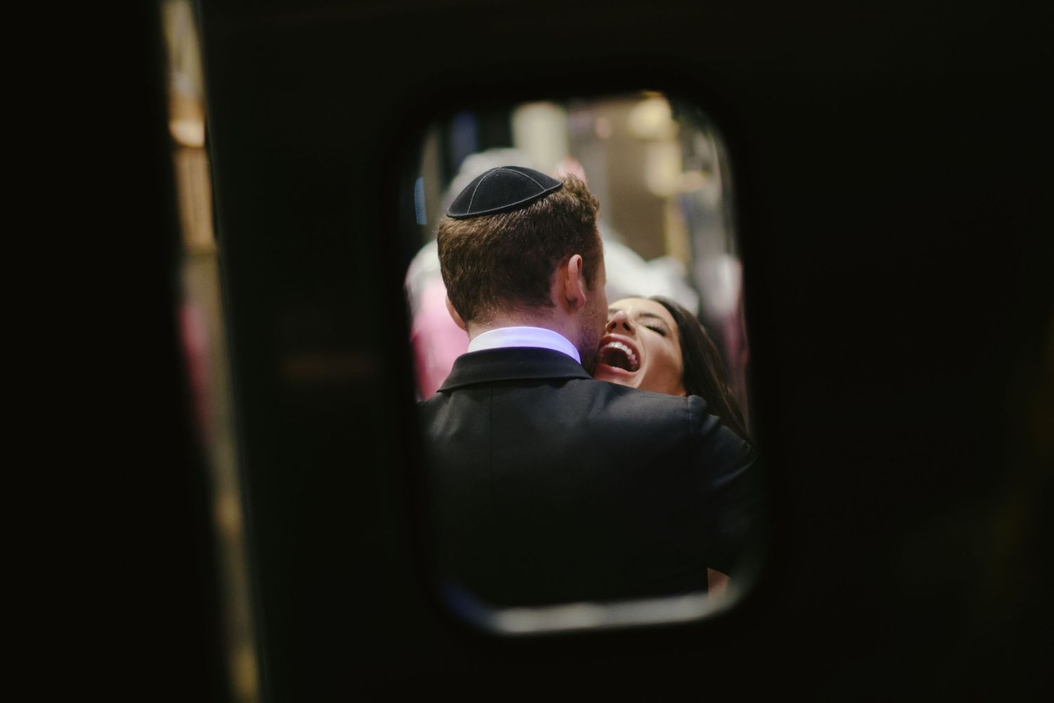 A candid wedding photo taken through a doorway showing a couple sharing an intimate moment at a Jewish ceremony.