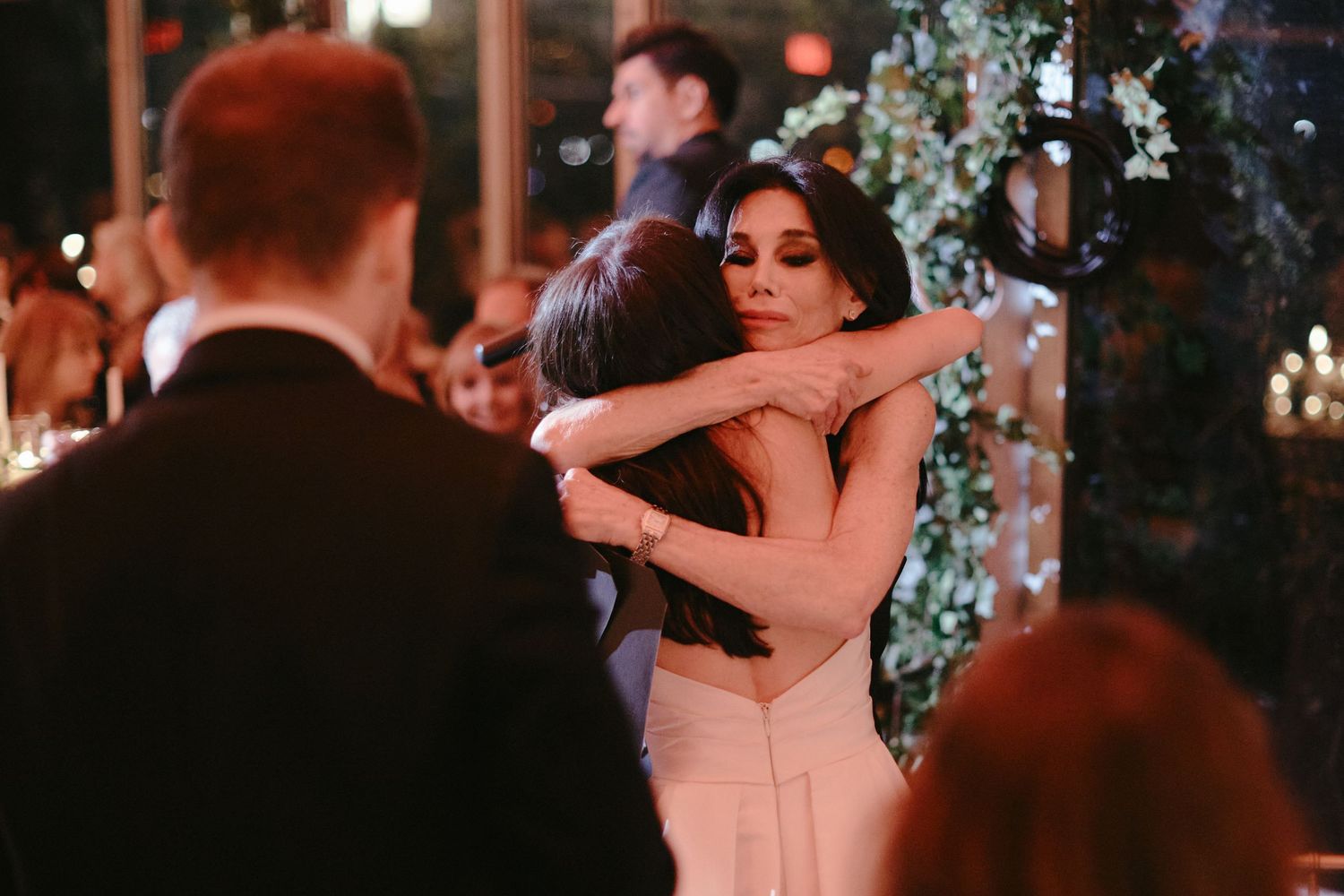People share an intimate dance embrace at an elegant evening reception with twinkling lights and floral decor in background.