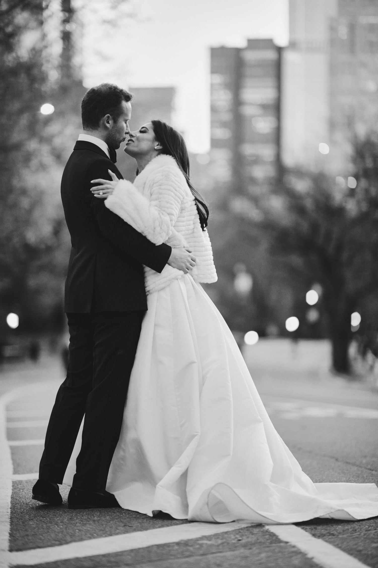 A romantic black and white wedding photograph shows a couple sharing a tender kiss on a city street at dusk.
