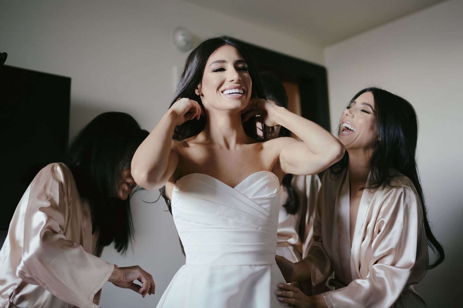 A joyful bride shares a candid moment with her bridesmaids in blush robes while getting ready before the ceremony.