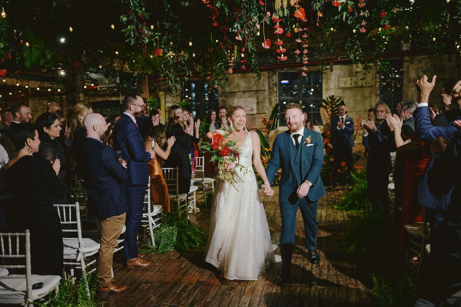 A wedding celebration under twinkling lights at a rustic outdoor venue with guests cheering along a brick pathway.