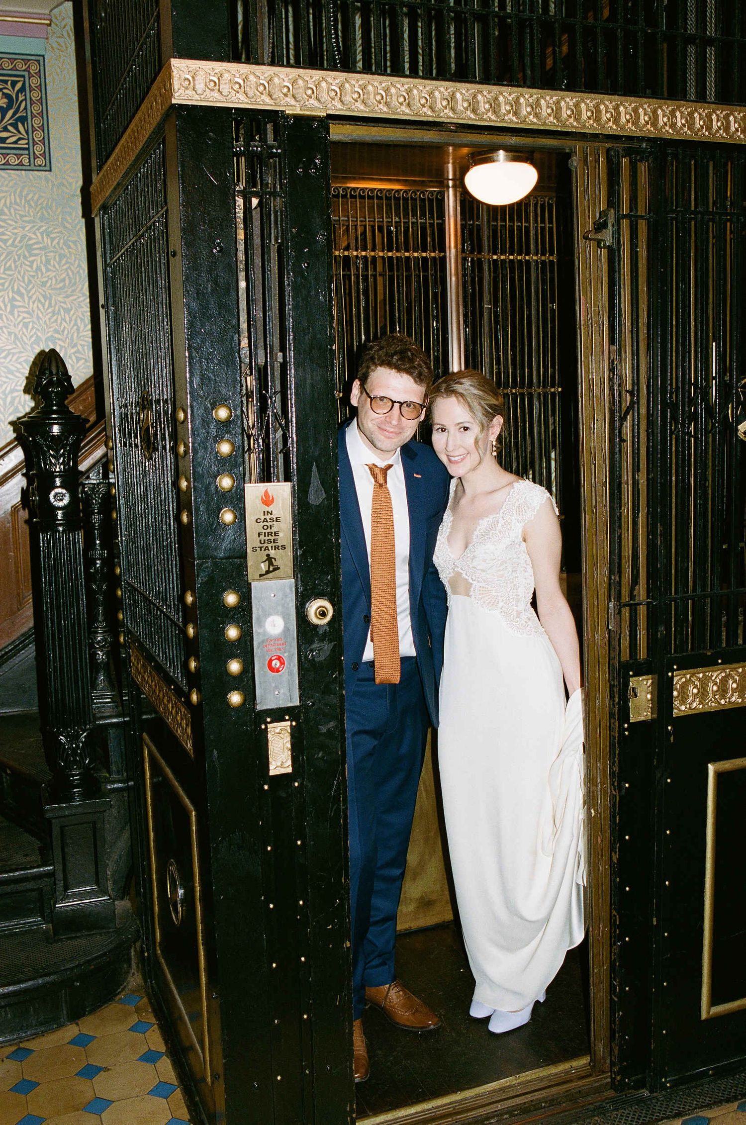 A pair of stylishly dressed people pose inside an ornate vintage elevator with dark wood paneling and decorative details.