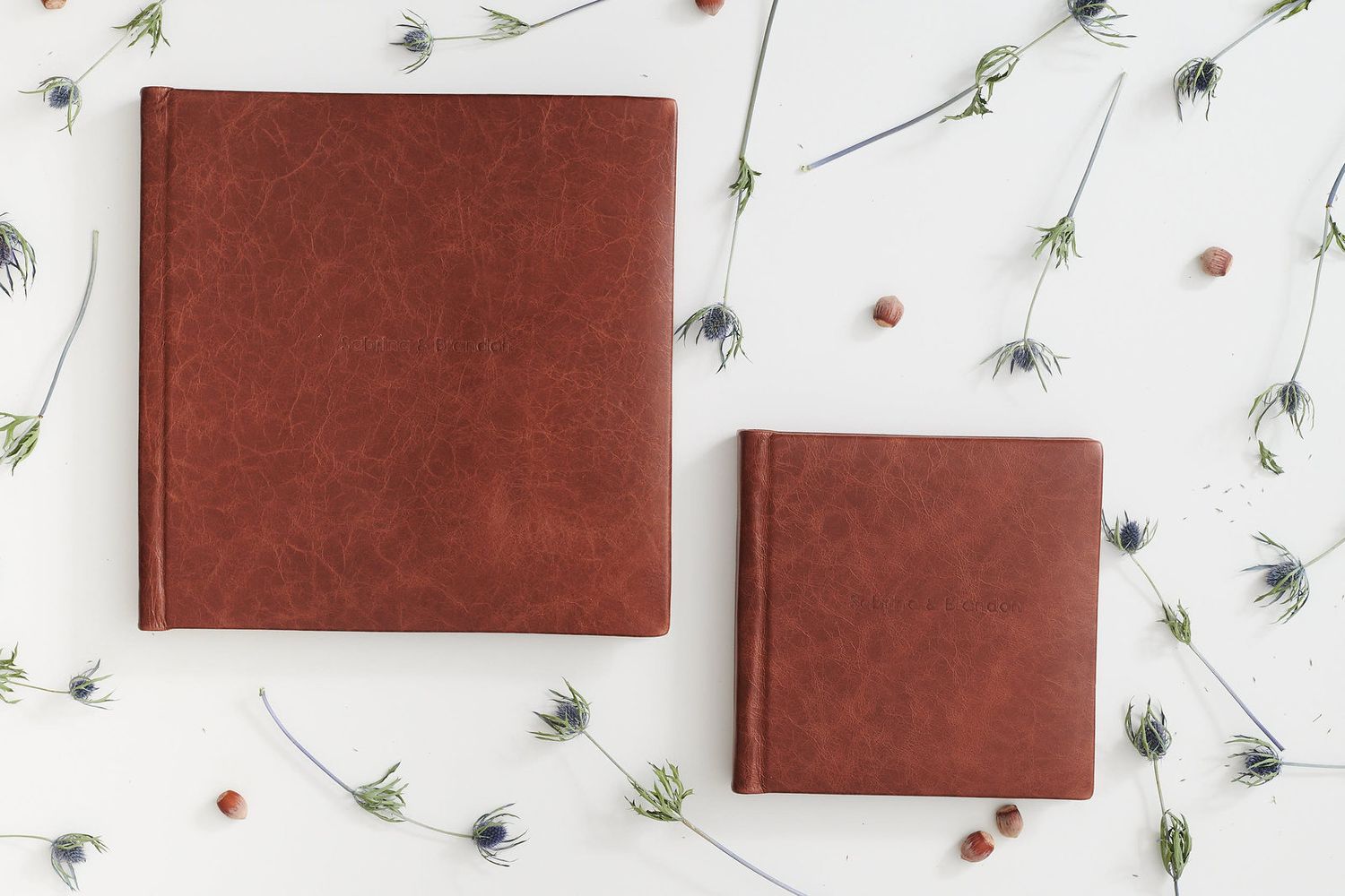 Two brown leather photo albums arranged on white surface with scattered small flowers and stems around them.