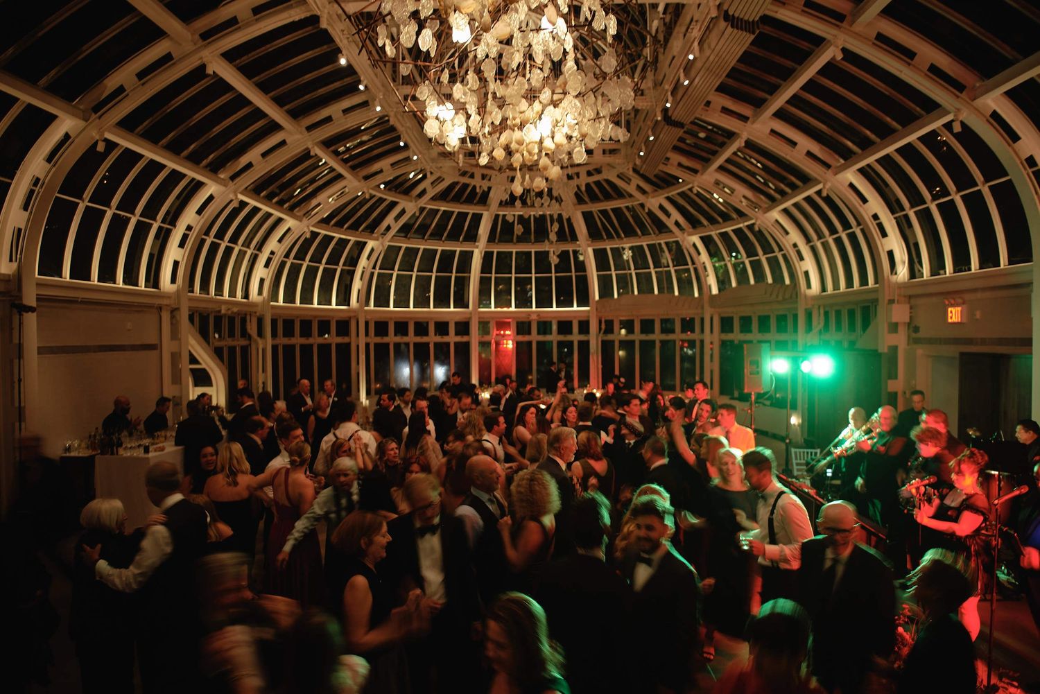 A large crowd dances beneath an ornate glass dome ceiling with glittering chandeliers and colorful party lighting.