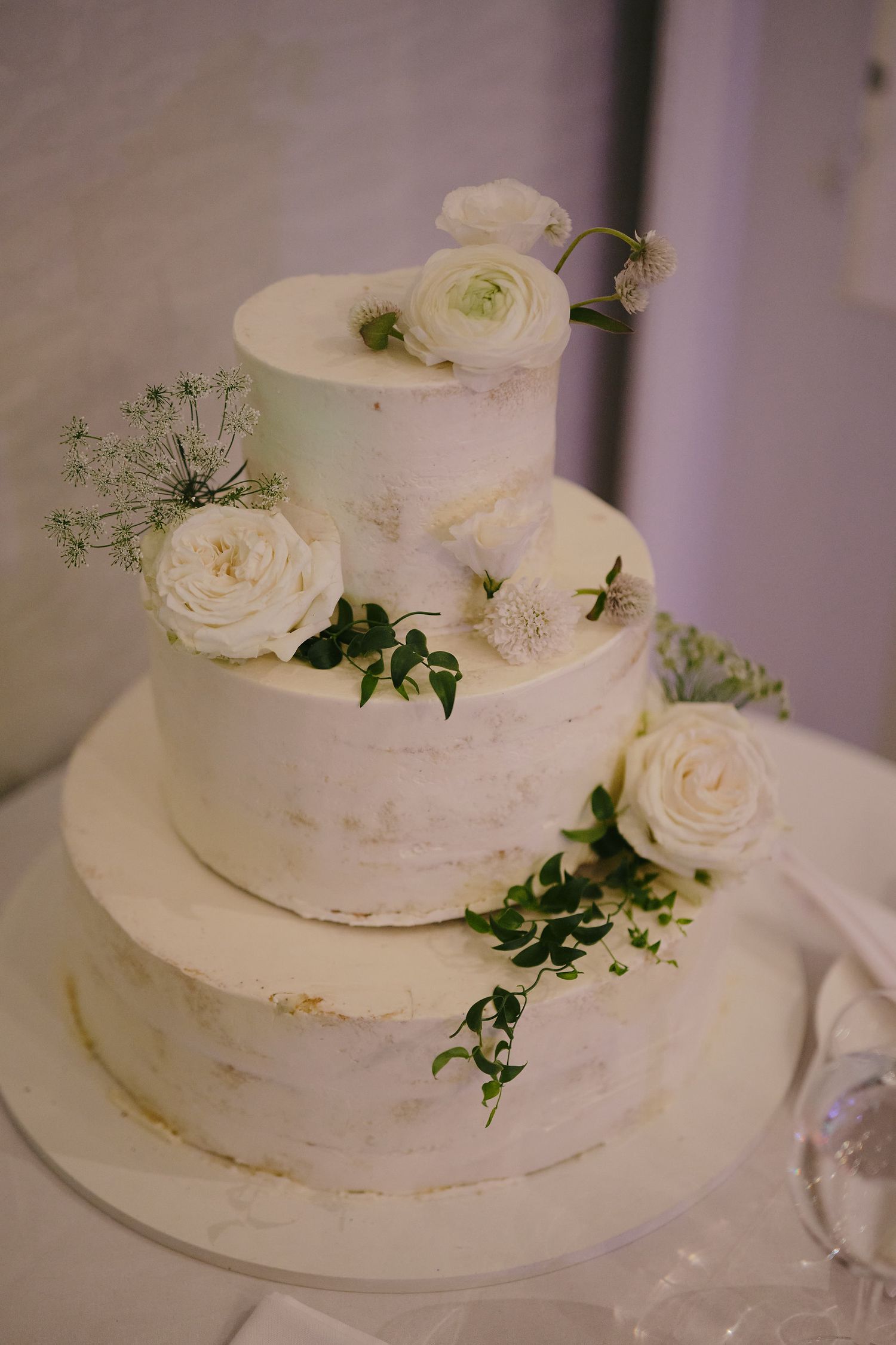 A three-tier white wedding cake decorated with fresh white roses and delicate greenery in a semi-naked style.