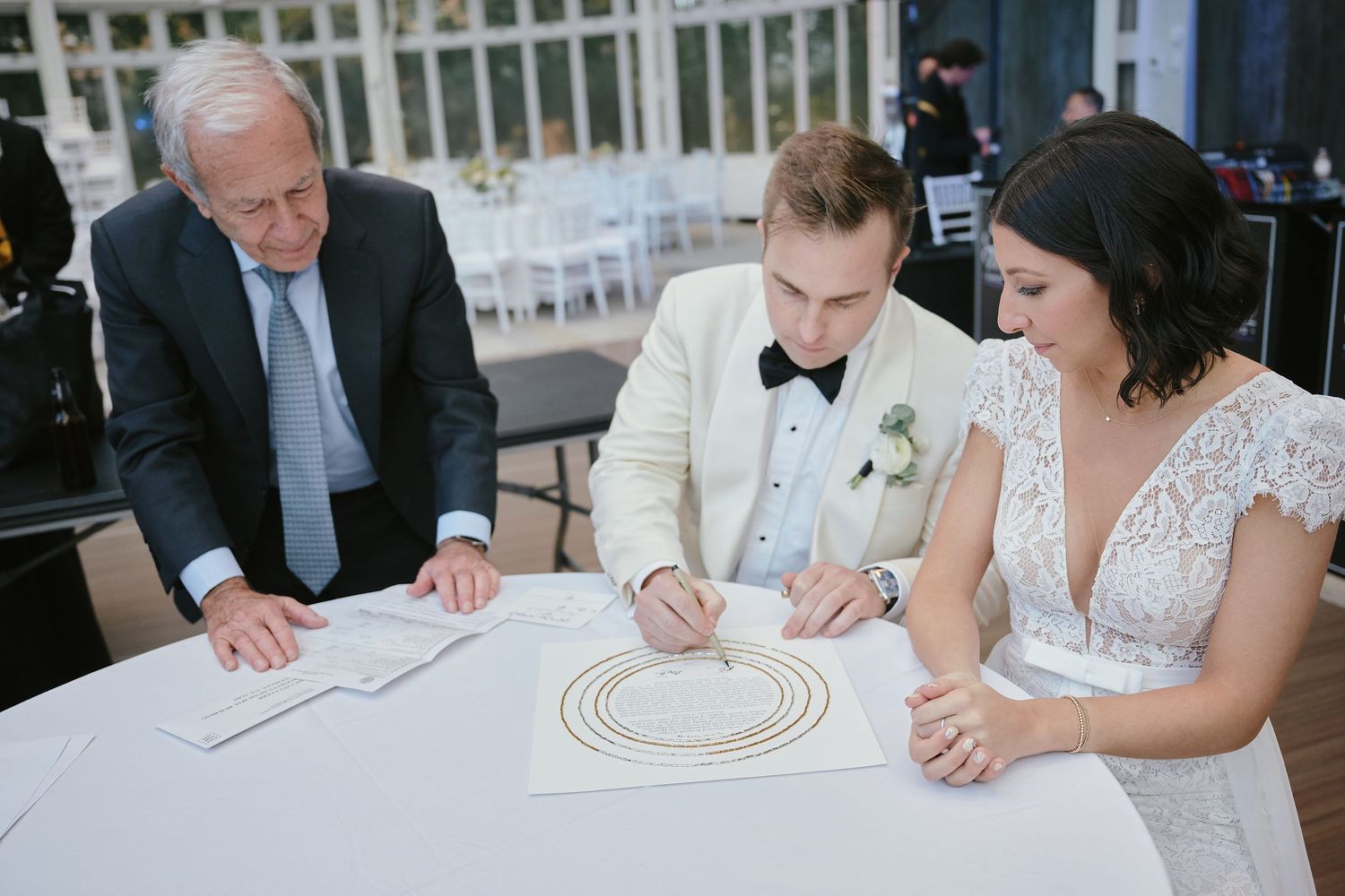 Couple signing documents at a round table during a wedding ceremony with an officiant present.