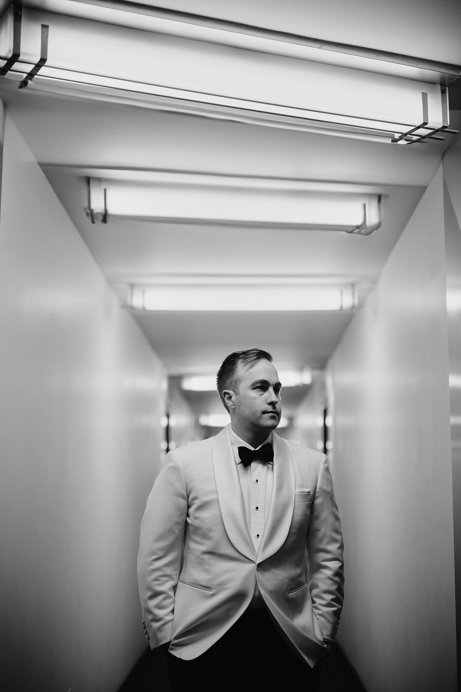 A black and white image of someone in a formal white tuxedo jacket and bowtie standing in a narrow hallway.