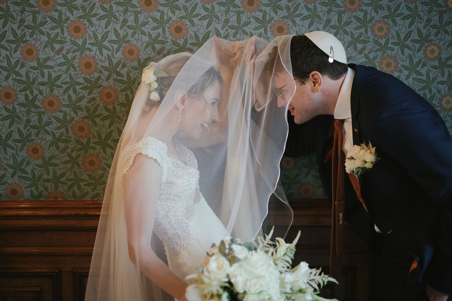 Bride and groom share an intimate moment under a flowing veil, with bride holding white bouquet against floral wallpaper.