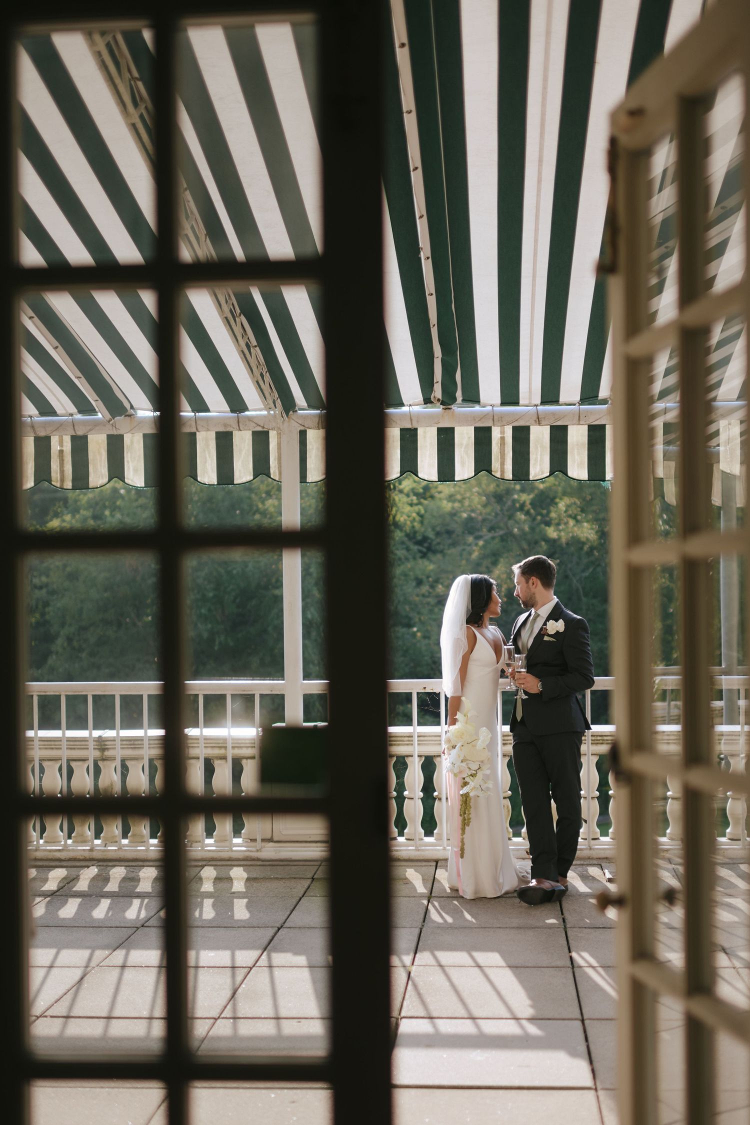 Bride and groom share a romantic moment on a striped awning balcony, framed by elegant French doors.