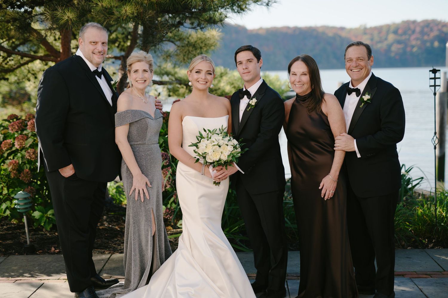 Wedding party poses outdoors near waterfront, bride in white gown holding bouquet, guests in formal black and brown attire.