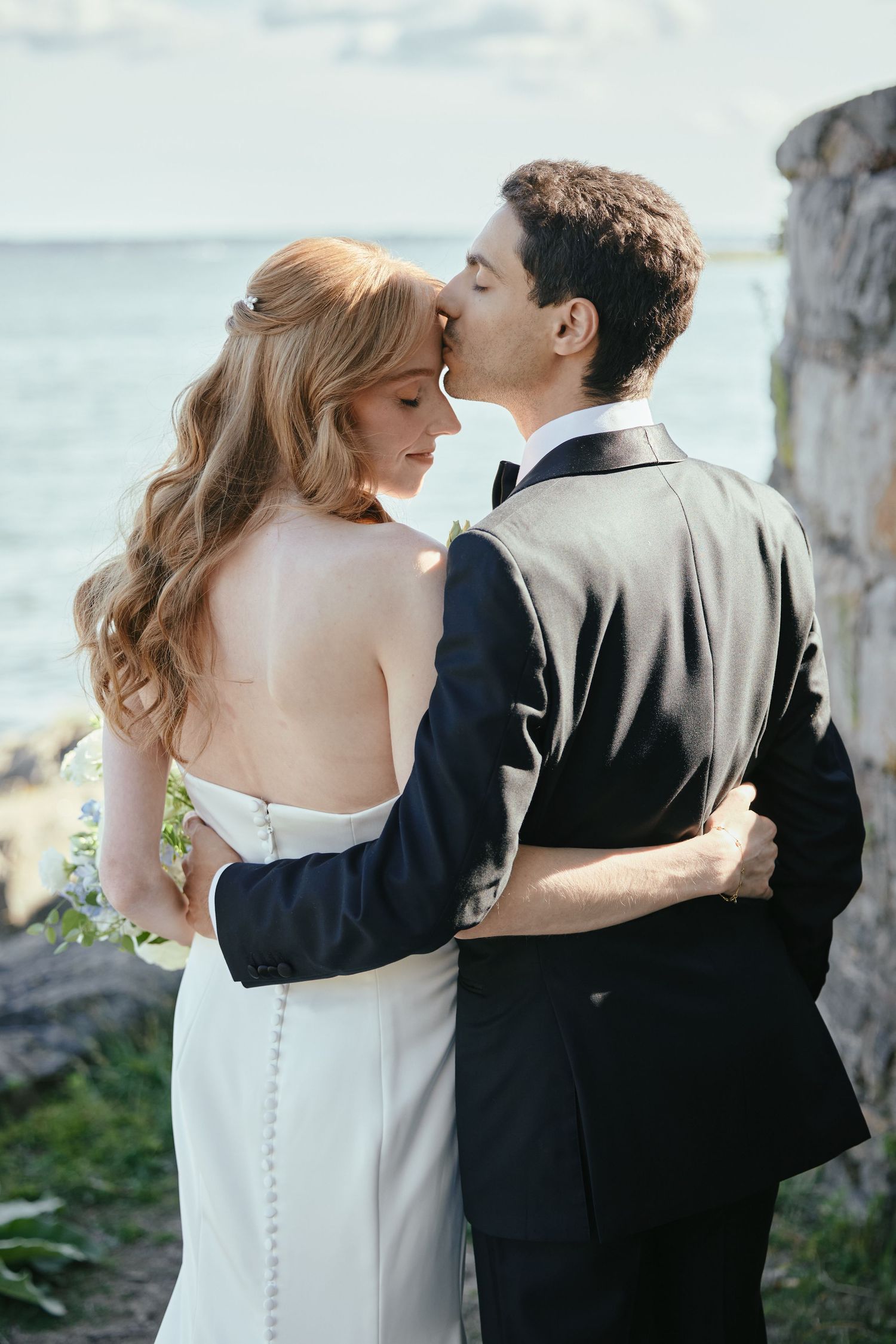A bride and groom share a tender kiss outdoors near the water, dressed in elegant wedding attire.