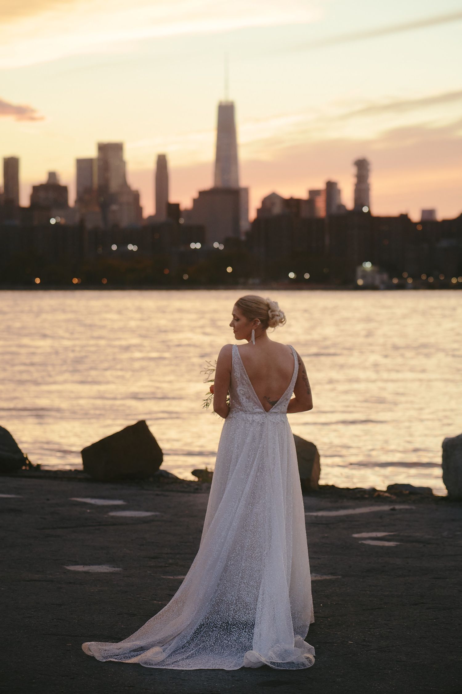 A white wedding dress silhouette stands against a dramatic city skyline at sunset by the waterfront.