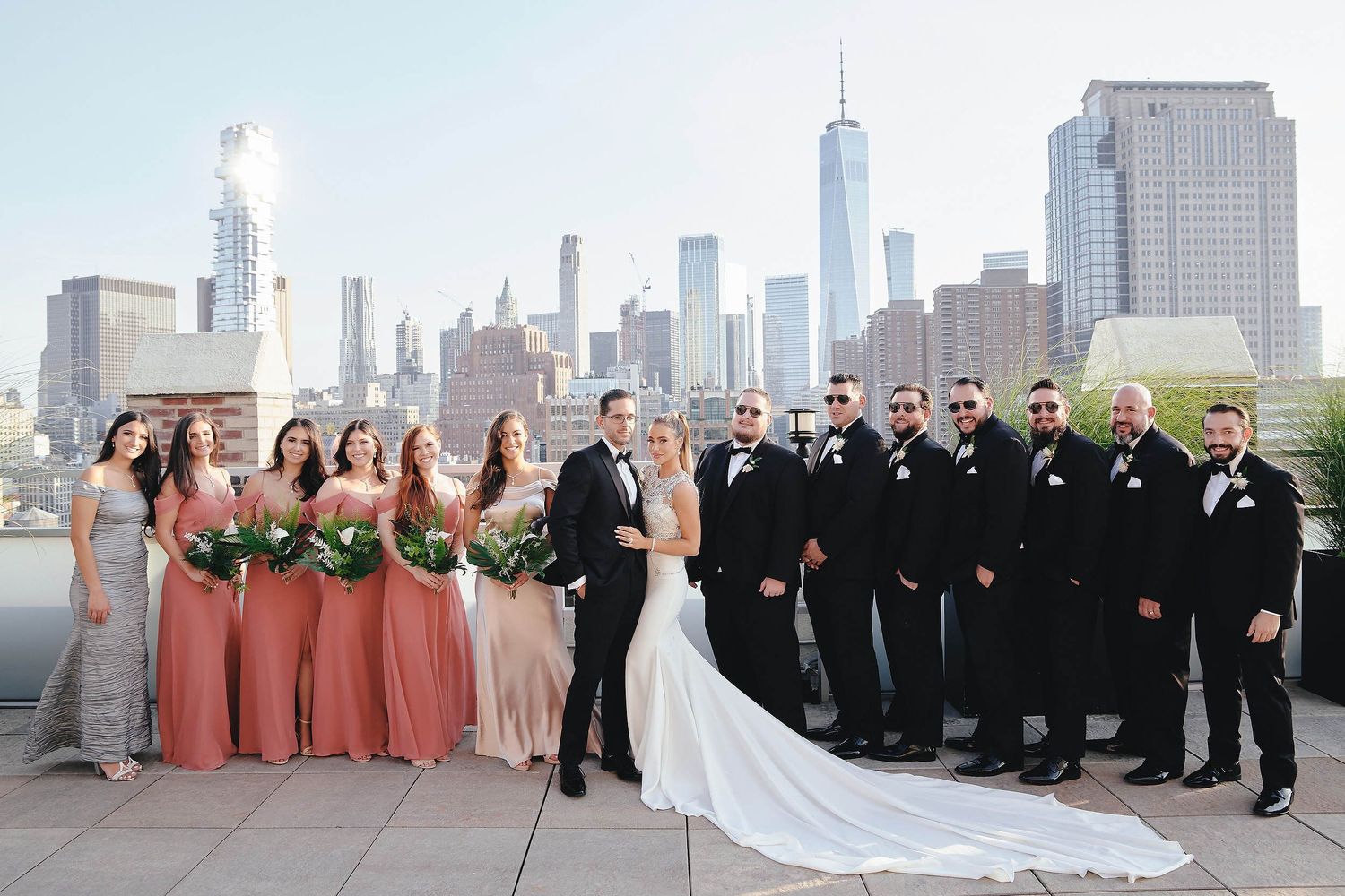Wedding party poses against the New York City skyline on a rooftop with One World Trade Center visible in the background.
