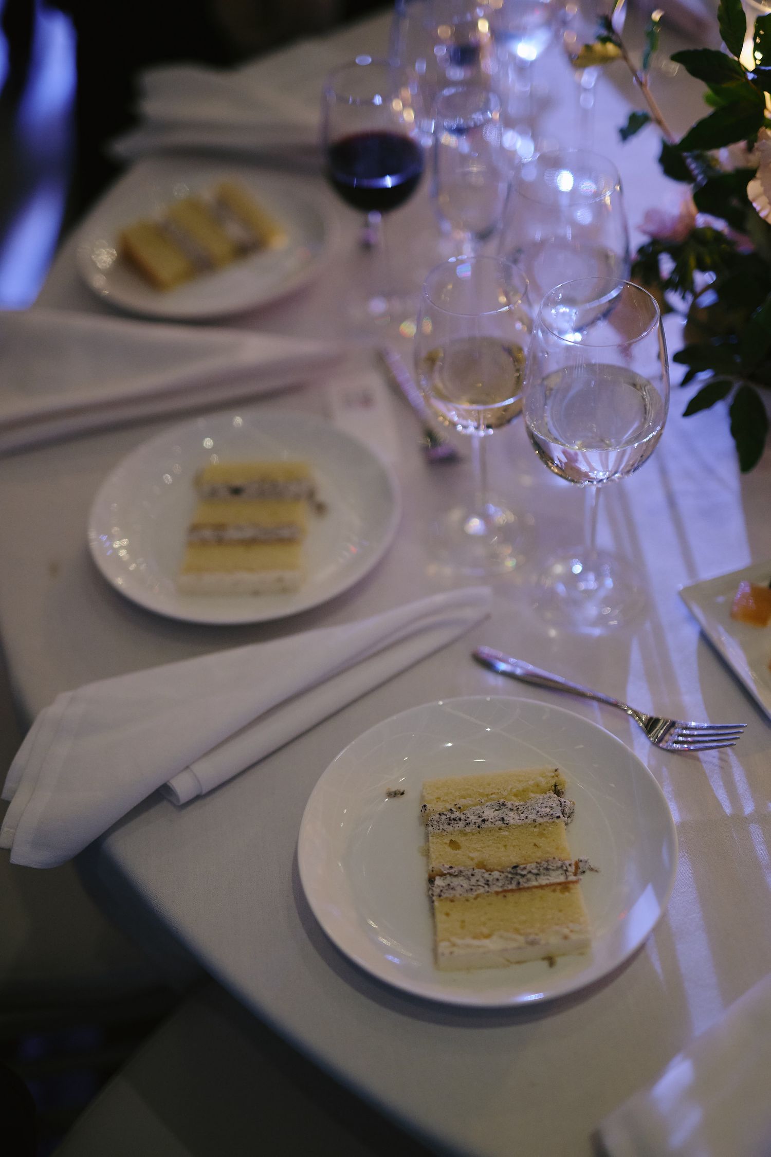 Elegant table setting with slices of layered white cake on plates alongside wine glasses and white linens.