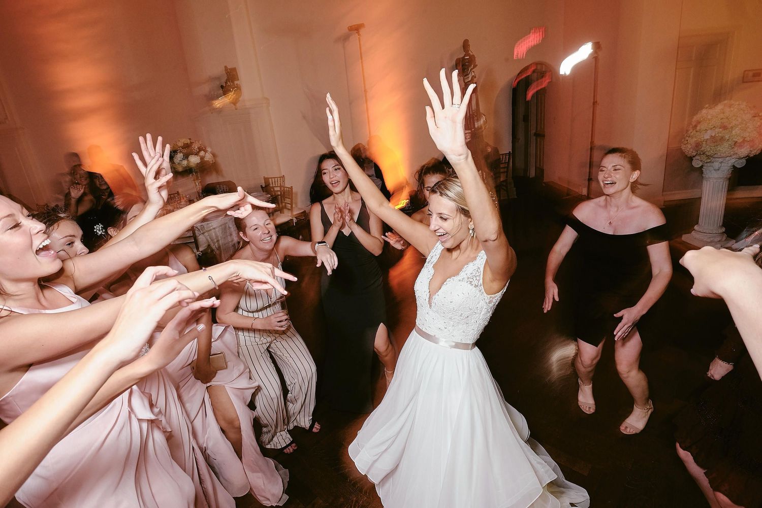 Wedding guests dance enthusiastically with arms raised on a dimly lit dance floor during the reception celebration.