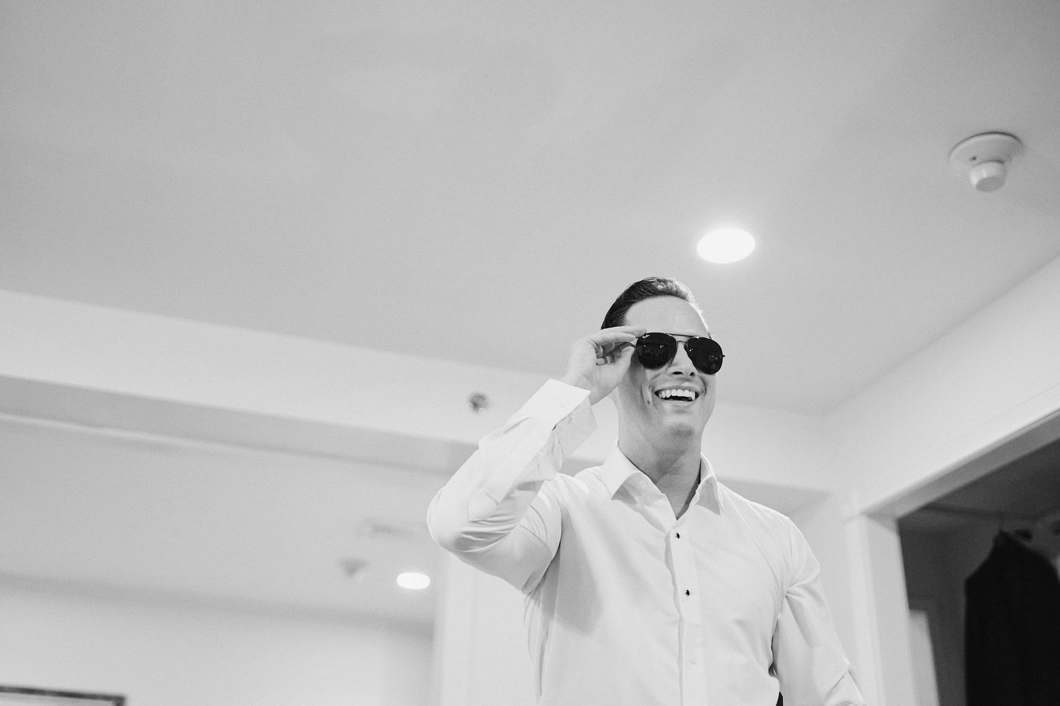 Black and white photo of someone wearing sunglasses and a white shirt smiling indoors.