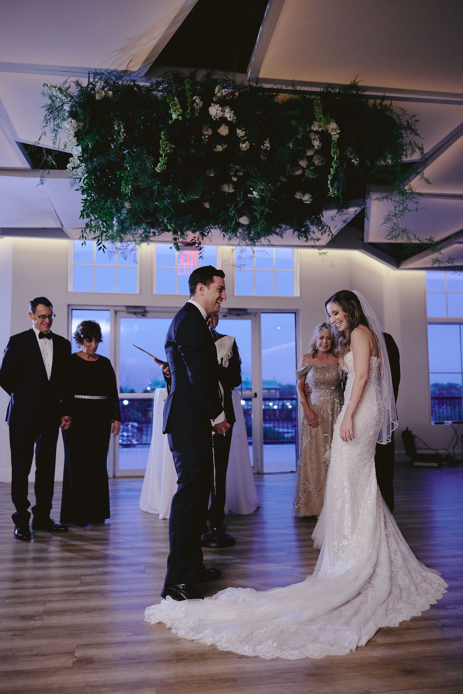 A bride in a lace wedding gown with a dramatic train stands in an event space with overhead greenery decorations.