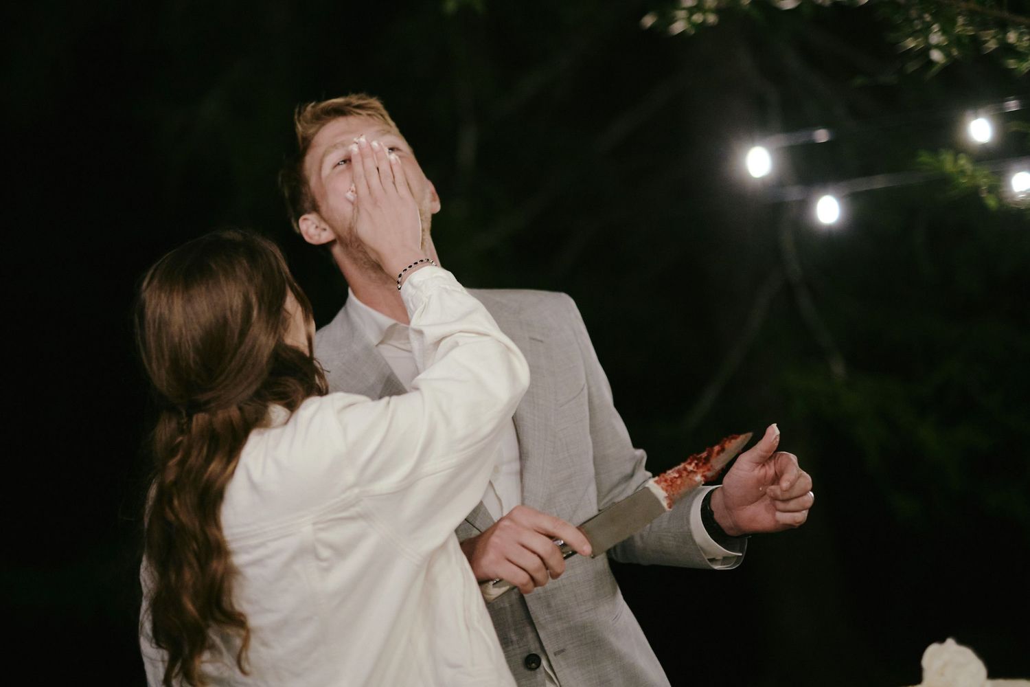 A couple shares a playful moment during their outdoor evening wedding celebration with string lights glowing in background.