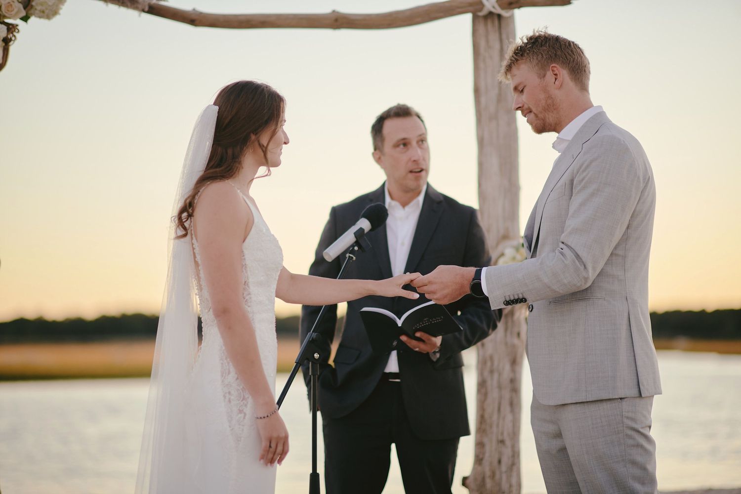 A wedding ceremony takes place at sunset by a lake with an officiant and couple under a rustic wooden arch.