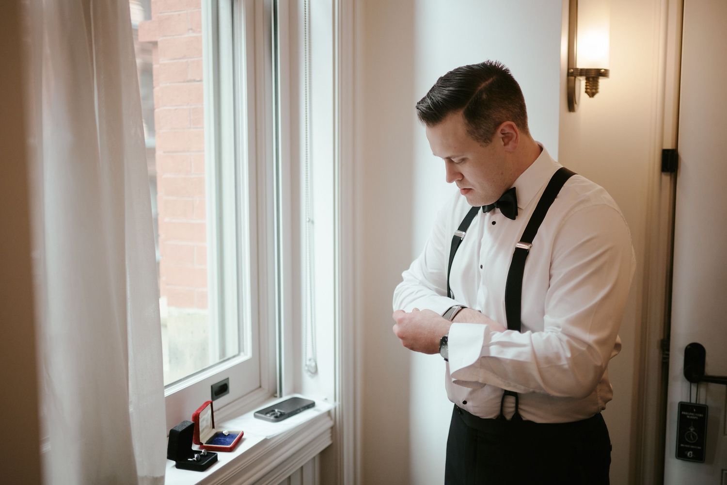 Groom in white shirt and suspenders adjusts his cuffs while standing by a window.