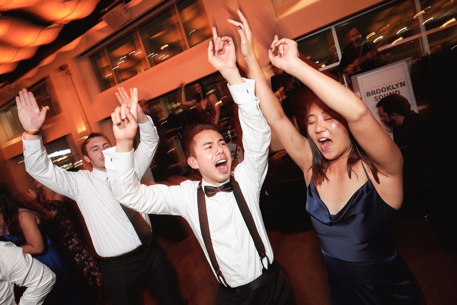 People dancing with their hands in the air at a celebration event, captured with warm orange lighting.